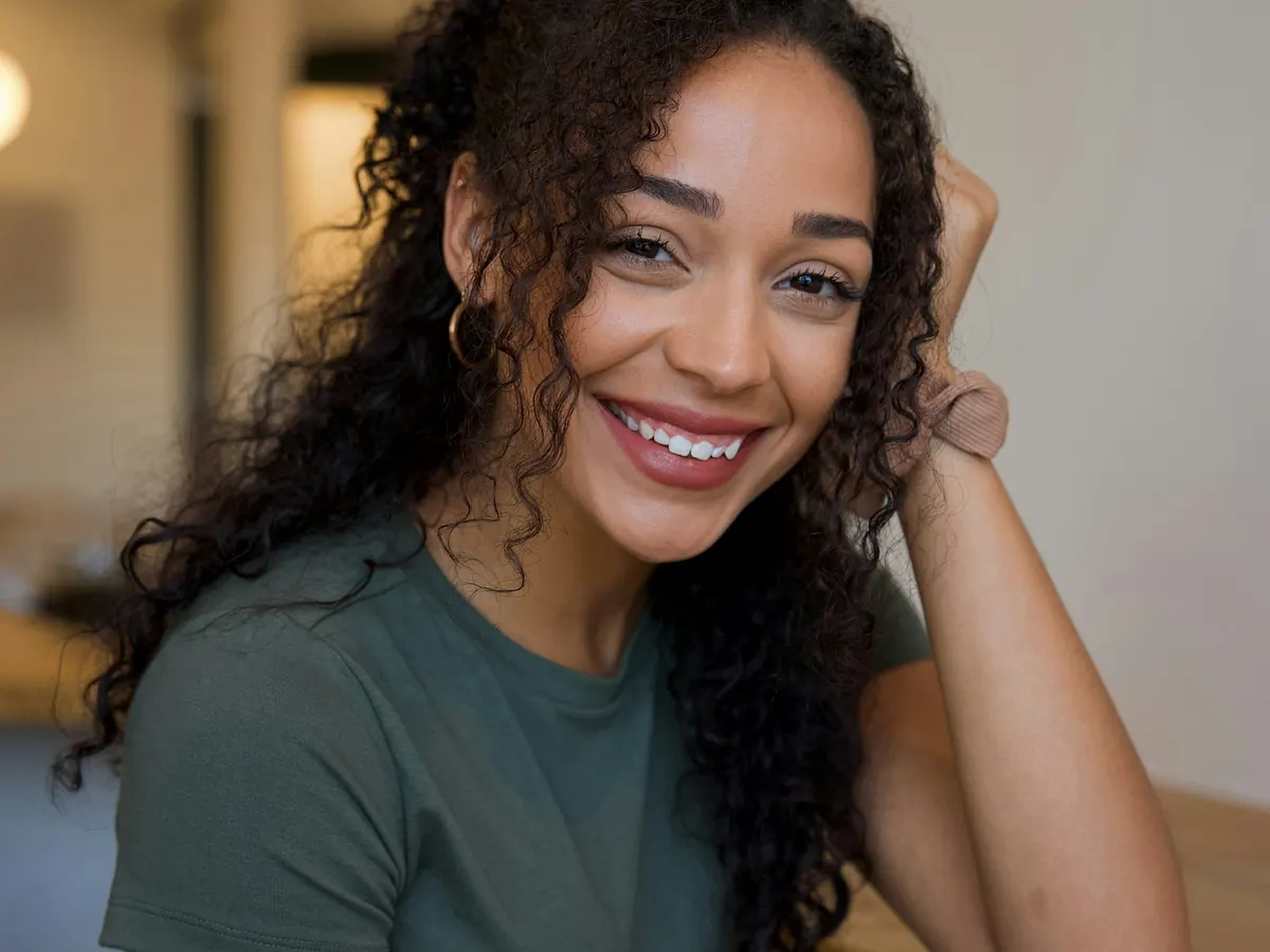 A smiling woman with curly hair sitting at a wooden table in natural light